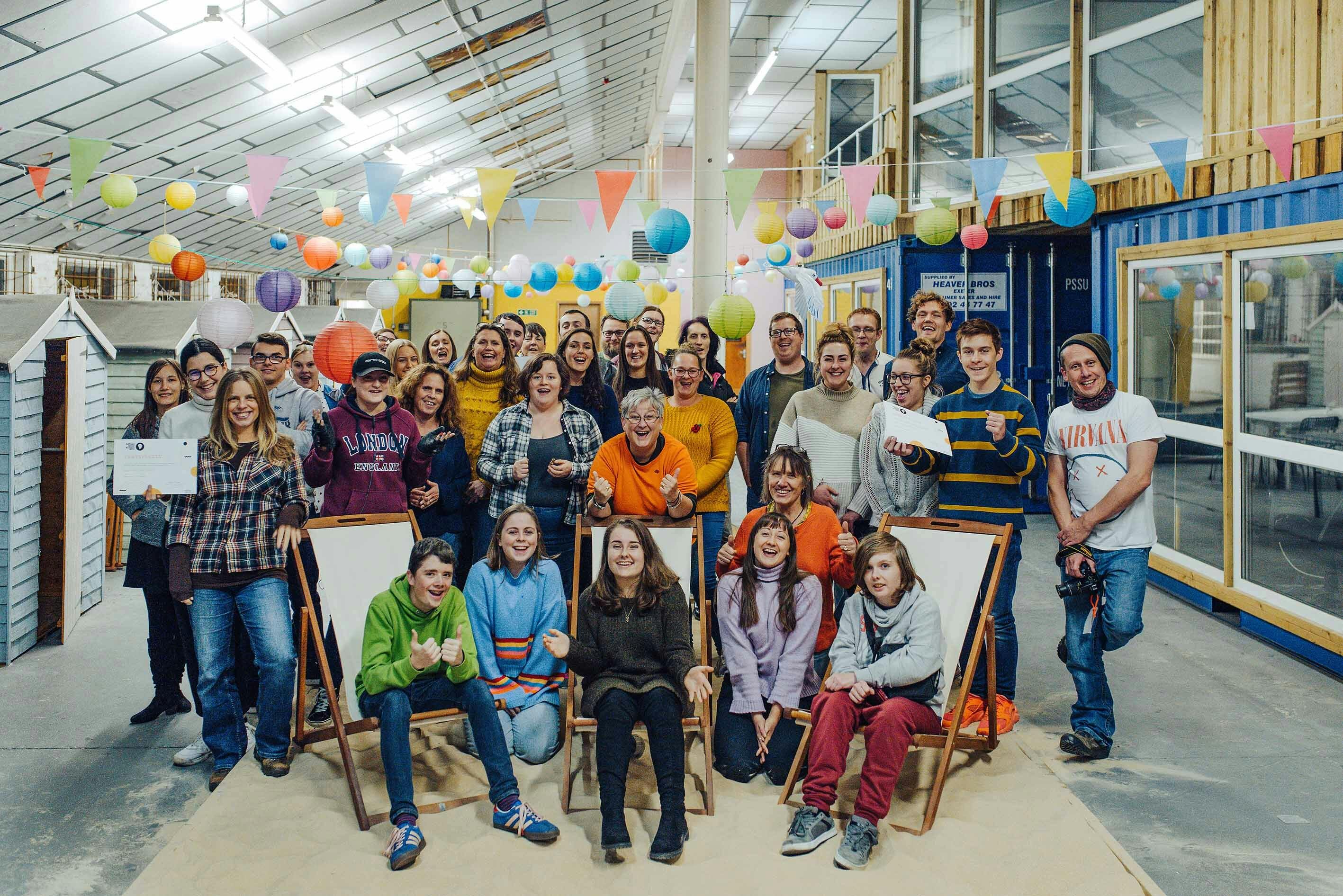 A large group of people pose for a photograph in a large warehouse style room strung with colourful bunting and paper lanterns AYCH Creative Jam November 2019