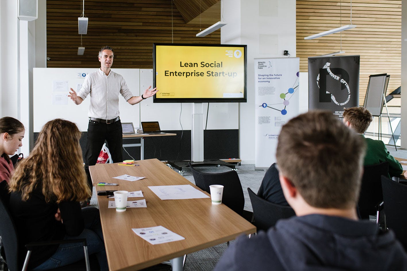 A man standing next to a screen showing a yellow slide talks to a group of people sat around a desk AYCH Hackfest June 2019