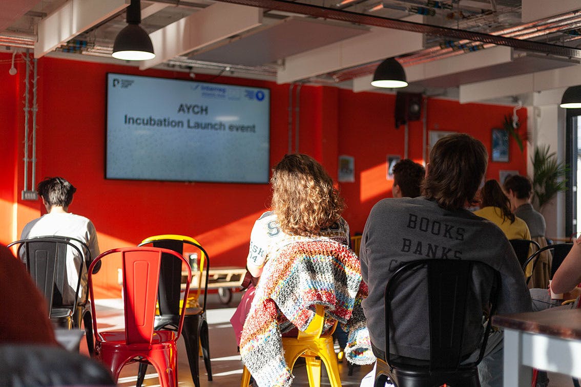 People sit on colourful chairs facing a screen on an orange wall AYCH Incubation Launch Event January 2020